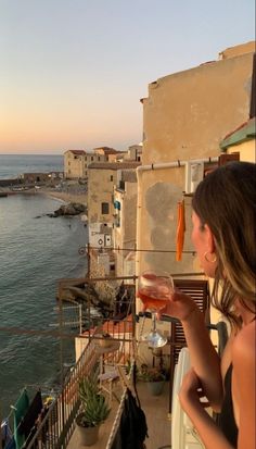 a woman holding a wine glass looking out at the ocean and buildings in the distance