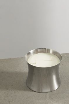 a white candle sitting in a metal bowl on top of a cement floor next to a wall