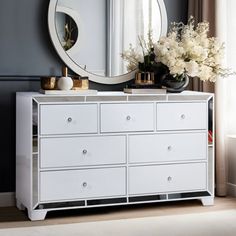 a white dresser with flowers and a round mirror on the wall above it in a room