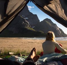 a woman sitting on top of a bed under a tent next to the ocean with mountains in the background