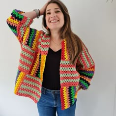 a woman wearing a colorful crocheted cardigan and jeans, standing in front of a white wall