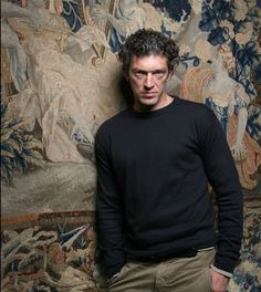 a man standing in front of a wall with tapestry behind him and looking at the camera