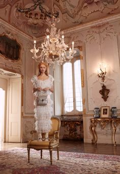 a woman in a white dress standing next to a chandelier and an antique chair