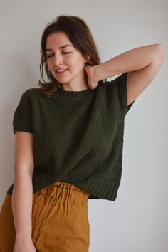 a woman wearing a green sweater and mustard colored pants with her hands behind her head