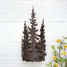 a metal pine tree on a white wooden wall next to yellow flowers and a potted plant