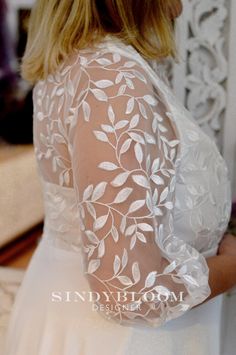 the back of a woman's white wedding dress with leaves on it and sheer sleeves