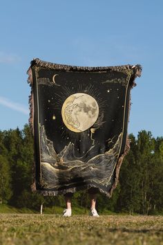 a person standing in the grass holding up a large black and gold blanket with an image of the moon on it