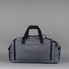 a grey duffle bag with black handles and straps on the handle, sitting against a gray background