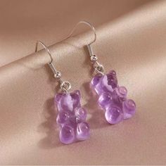 a pair of purple teddy bear earrings sitting on top of a pink satin material surface