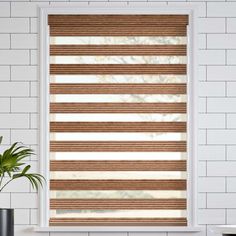 a white brick wall with a window covered in brown and beige shades, next to a potted plant