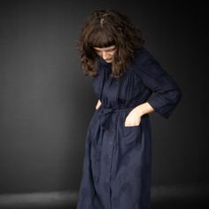 a woman standing in front of a black background wearing a long blue dress with pockets