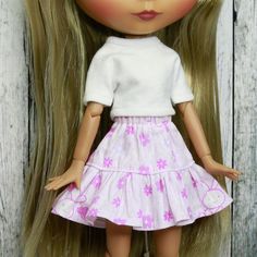 a doll with long blonde hair wearing a white shirt and pink flowered skirt, standing next to a wooden fence