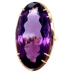 This large enchanting 14.50 carat faceted elongated oval amethyst is set in a mid-century modern 14 karat yellow gold setting that has a rose gold hue. This amethyst's rich deep purple comes alive in the light enhancing its brightness. The stone measures approximately 1 1/16" long by 1/2" wide and is held in the mounting by four stations of double prongs. Under the gem, the mounting appears as a frame highlighting the beauty of the stone in this circa 1970 mounting making the top of the ring approximately 1 1/4" long by 5/8" wide. This vibrant amethyst is very clean with no visible inclusions. The ring rises approximately 7/16" off the finger, is a US size 6.5, and weighs 9.1 grams. The 1970's was a decade of punk rock and disco, social change, and iconic movies such as The Godfather, Star Luxury Formal Rings With Oval Cabochon, Luxury Formal Amethyst Ring Oval Cabochon, Luxury Purple Oval Cabochon Rings, Luxury Hallmarked Oval Cabochon Amethyst Ring, Luxury Hallmarked Amethyst Oval Cabochon Ring, Luxury Oval Amethyst Ring, Art Deco Style, Luxury Yellow Gold Amethyst Cabochon Ring, Luxury Oval Amethyst Ring In Art Deco Style, Luxury Vintage Amethyst Oval Cabochon Ring