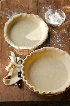 two pie crusts on a wooden table next to a knife and bowl with flour