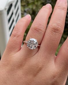 a woman's hand with a diamond ring on top of her finger, in front of some bushes