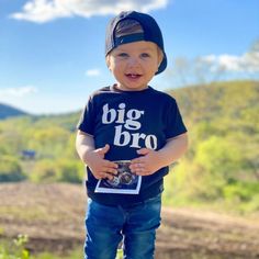 a little boy that is standing up with a camera in his hand and wearing a big bro t - shirt