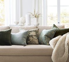 a couch with many pillows on it in front of a white wall and large windows