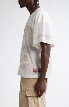 a man with dreadlocks standing in front of a white wall wearing a white shirt and khaki shorts