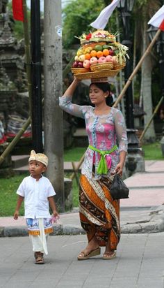 a woman carrying a basket on her head while walking down the street with a small boy