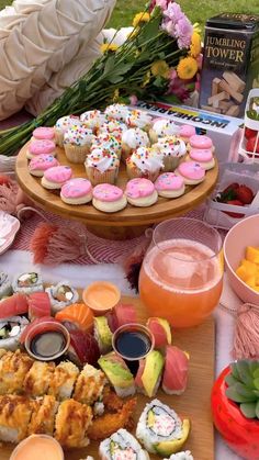 an assortment of sushi and cupcakes on a wooden platter with flowers in the background