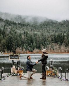 a man kneeling down next to a woman on a dock near a lake and trees