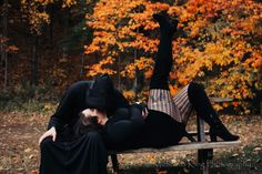 two people are laying on a bench in front of trees with orange and yellow leaves