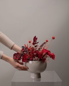 a woman is arranging flowers in a white vase on a clear pedestal with her hands