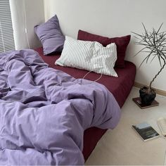 a bed with purple sheets and pillows in a bedroom next to a plant on the floor