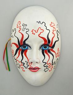 a white mask with red and black designs on it's face is hanging from a wall