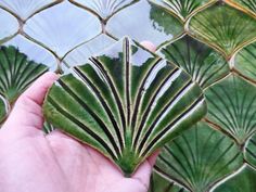 a hand holding a piece of green tile in front of a fence with leaves on it