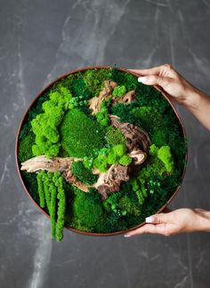 two hands holding a bowl filled with broccoli and other green vegetables on top of a table