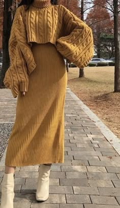 a woman is walking down the sidewalk wearing a mustard colored sweater dress and white boots