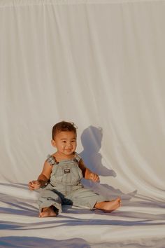 a baby sitting on the ground in front of a white backdrop smiling at the camera