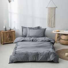 a bed with grey sheets and pillows in a room next to a wooden table on the floor