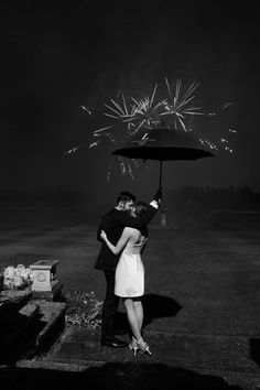 black and white photograph of couple kissing under an umbrella with fireworks in the sky behind them