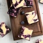 blueberry cheesecake bars on a wooden cutting board with fresh blueberries around them