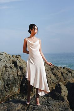 a woman in a white dress standing on rocks near the ocean with her hands behind her head