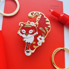 a red and white tiger pin sitting on top of a red book next to a pair of gold rings