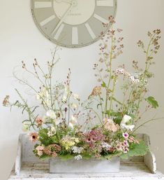 an arrangement of flowers in front of a large clock on the wall above a bench