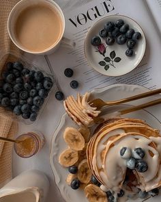 pancakes with bananas and blueberries on a plate next to cups of coffee, spoons and saucers