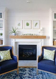 two blue chairs sitting in front of a fireplace with pictures on the wall behind them