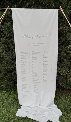 a white wedding seating chart hanging from a clothes line in front of some bushes and trees