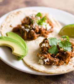 two tacos with meat and cilantro on a plate next to an avocado