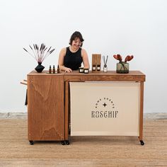 a woman standing behind a counter with bottles and candles on it, in front of a white wall