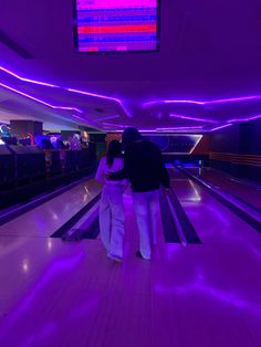 two people walking down a bowling alley with purple lights on the ceiling and television above them