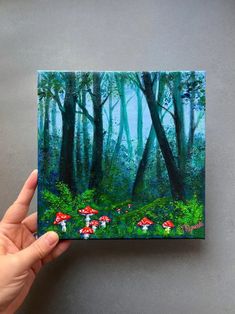 a hand holding up a small painting of mushrooms in the middle of a green forest