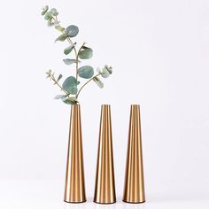 three gold vases with green leaves in them on a white surface, one is tall and the other is small