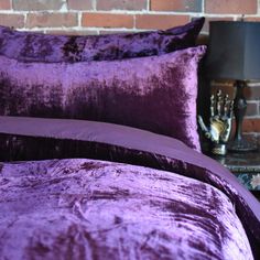 a bed with purple sheets and pillows in front of a brick wall on a night stand