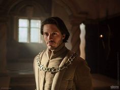 a man wearing a chain around his neck in a dark room with sunlight streaming through the window