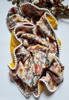 a yellow scarf with flowers and pom - poms on the bottom is laying next to an arrangement of dried flowers